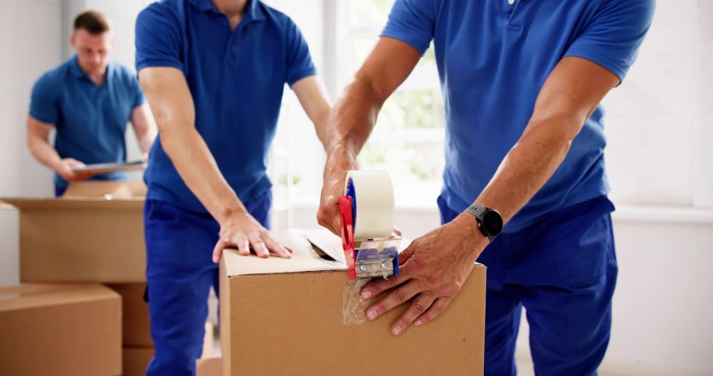 Close up shot of two movers taping up a box while another packs a box in the background.