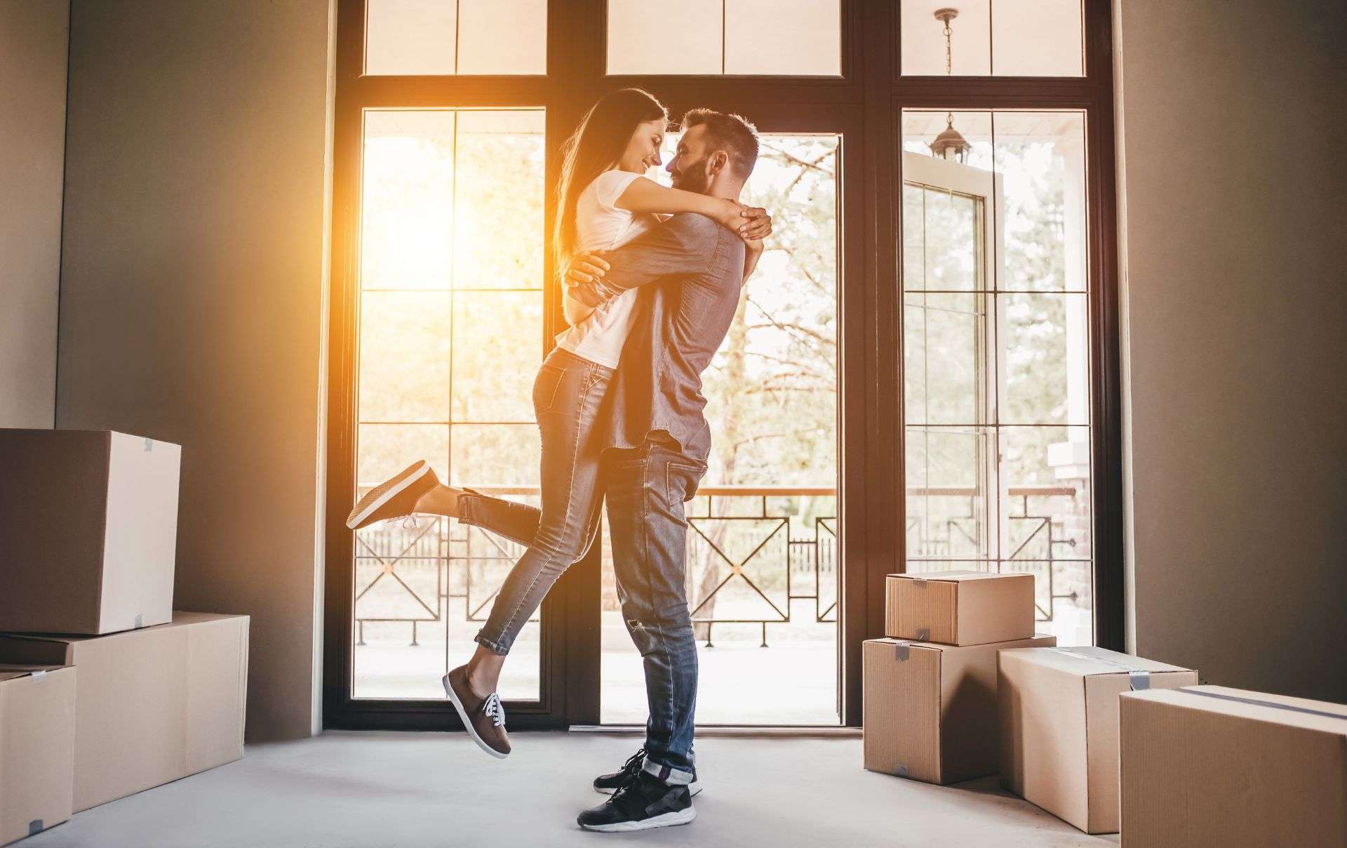 A man lifting a woman in a celebratory hug in the middle of a sunlit living room filled with cardboard moving boxes, with large glass doors leading to a balcony in the background.