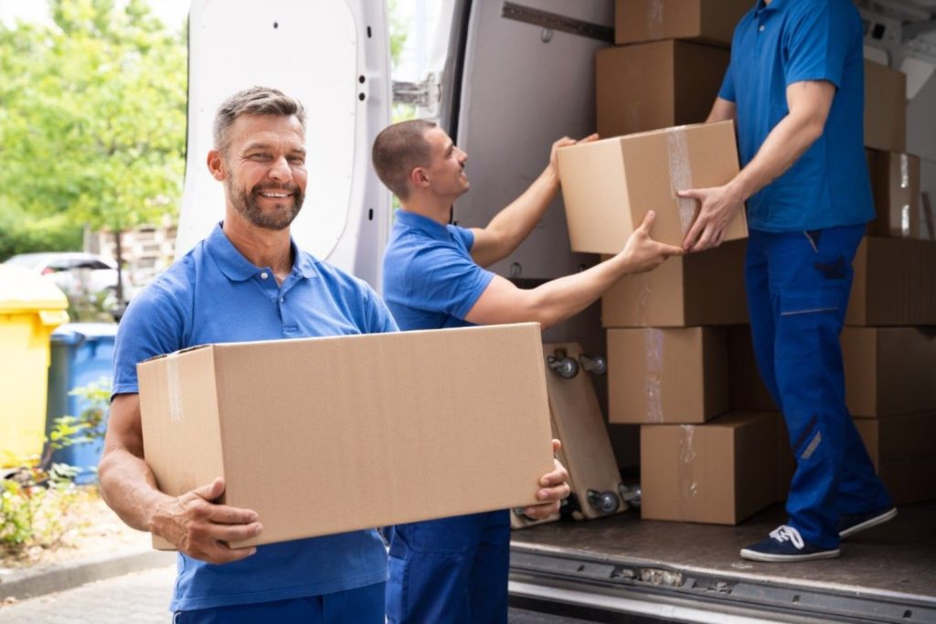 Mover smiling while holding a box while two other movers unload a moving truck in the background